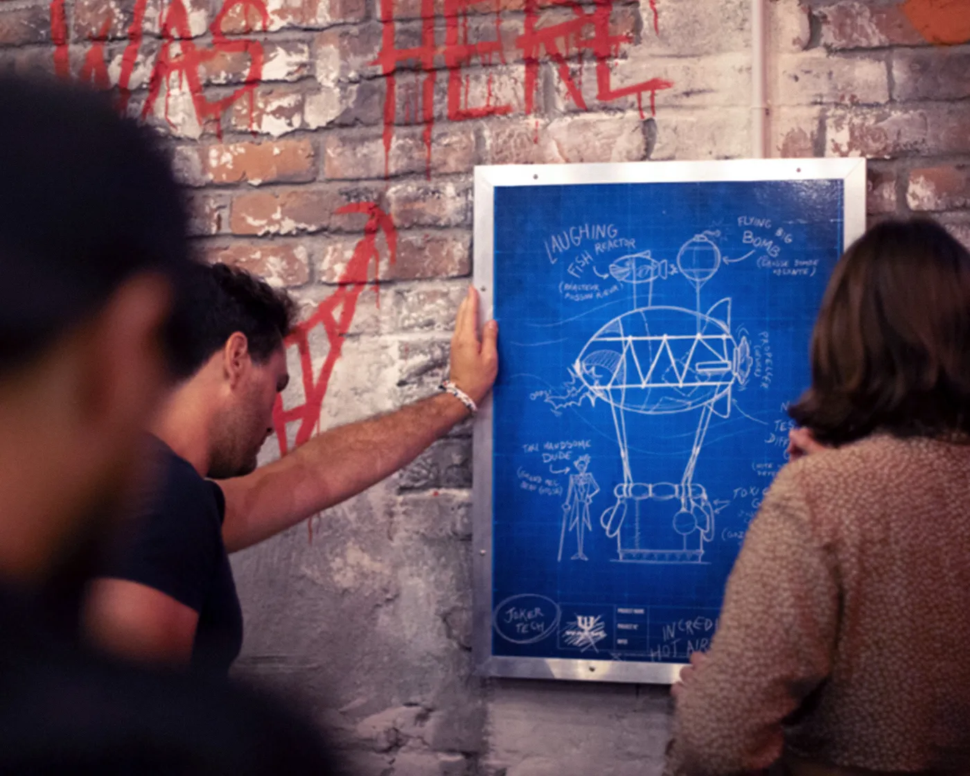 Visitors examining a blueprint poster of the Joker's flying-fish device on a graffitied wall