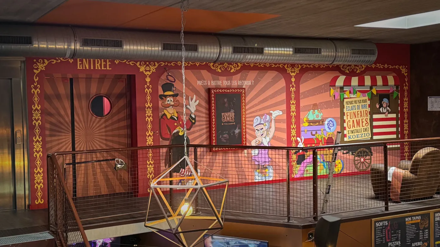 Funfair Games entrance at Koezio with circus-style murals of Archibald and Micheline and a fairground booth sign