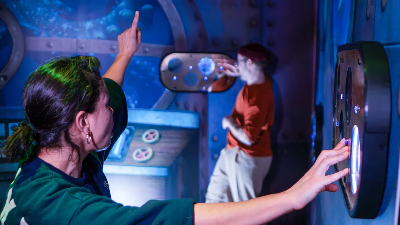 Two players touching illustrated wall panels and a porthole in the submarine set