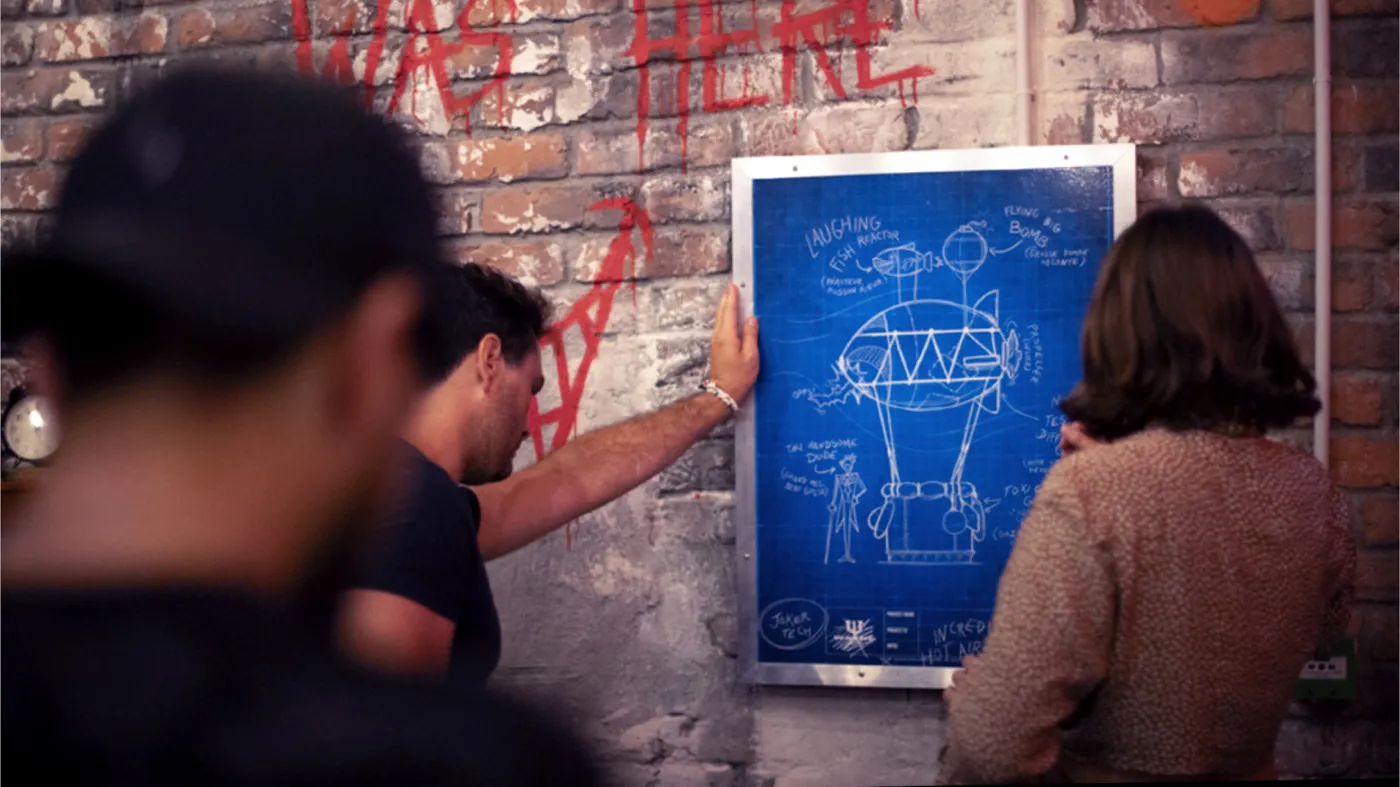 Deux personnes étudient le blueprint d'un engin du Joker affiché sur un mur de briques.