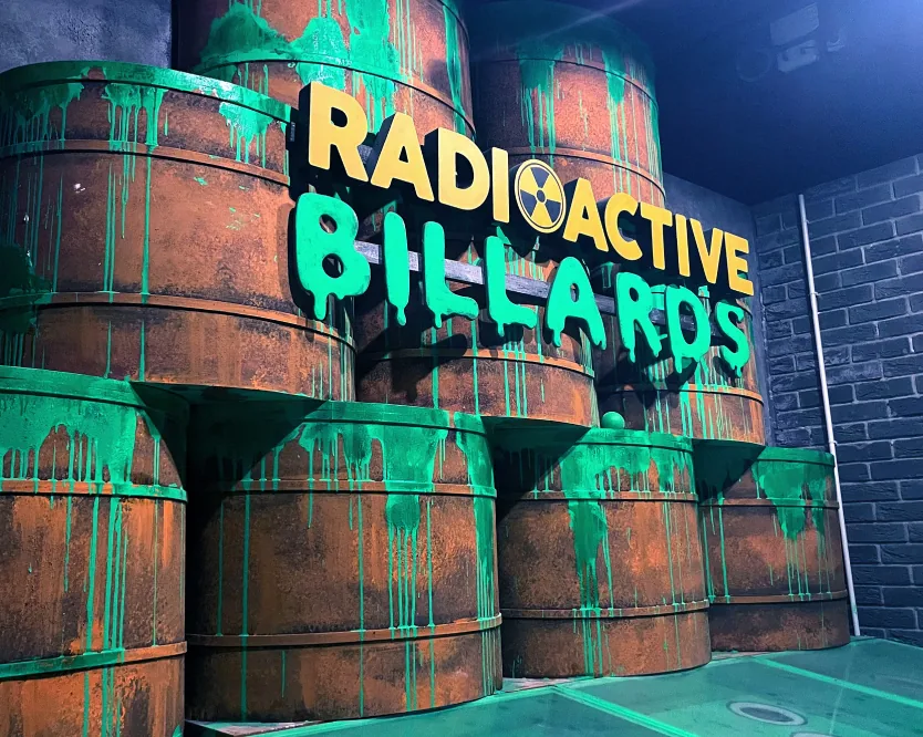 Enseigne néon « Radioactive Billiards » verte au-dessus de fûts toxiques empilés.