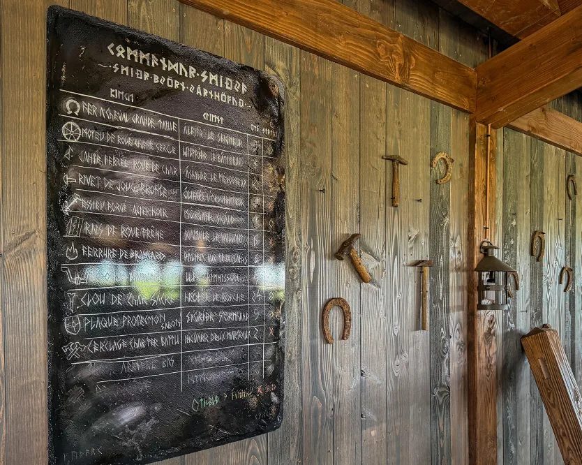 Menu sur ardoise aux runes, fixé à un mur lambrissé, avec un fer à cheval et des outils de forge.